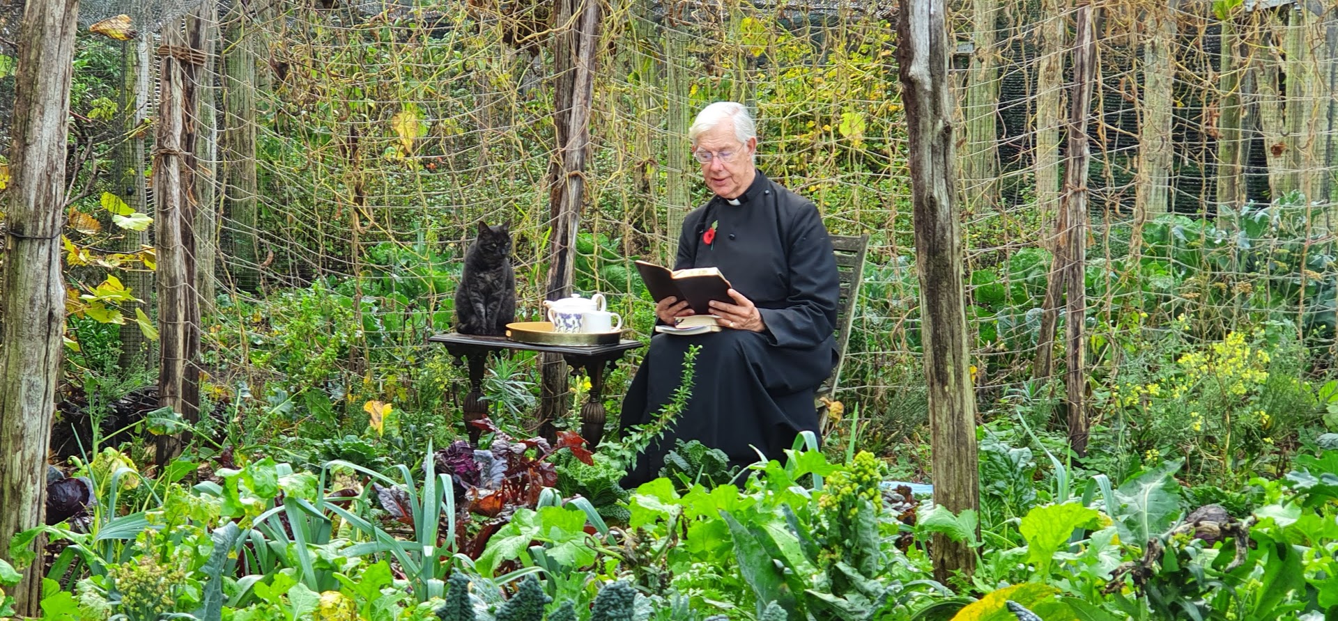 Dean Robert Willis sitting with cat Leo in the deanery garden reading from a Bible